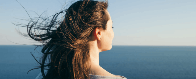 A woman looking out over the ocean, with her hair blowing in the wind beneath a clear blue sky.