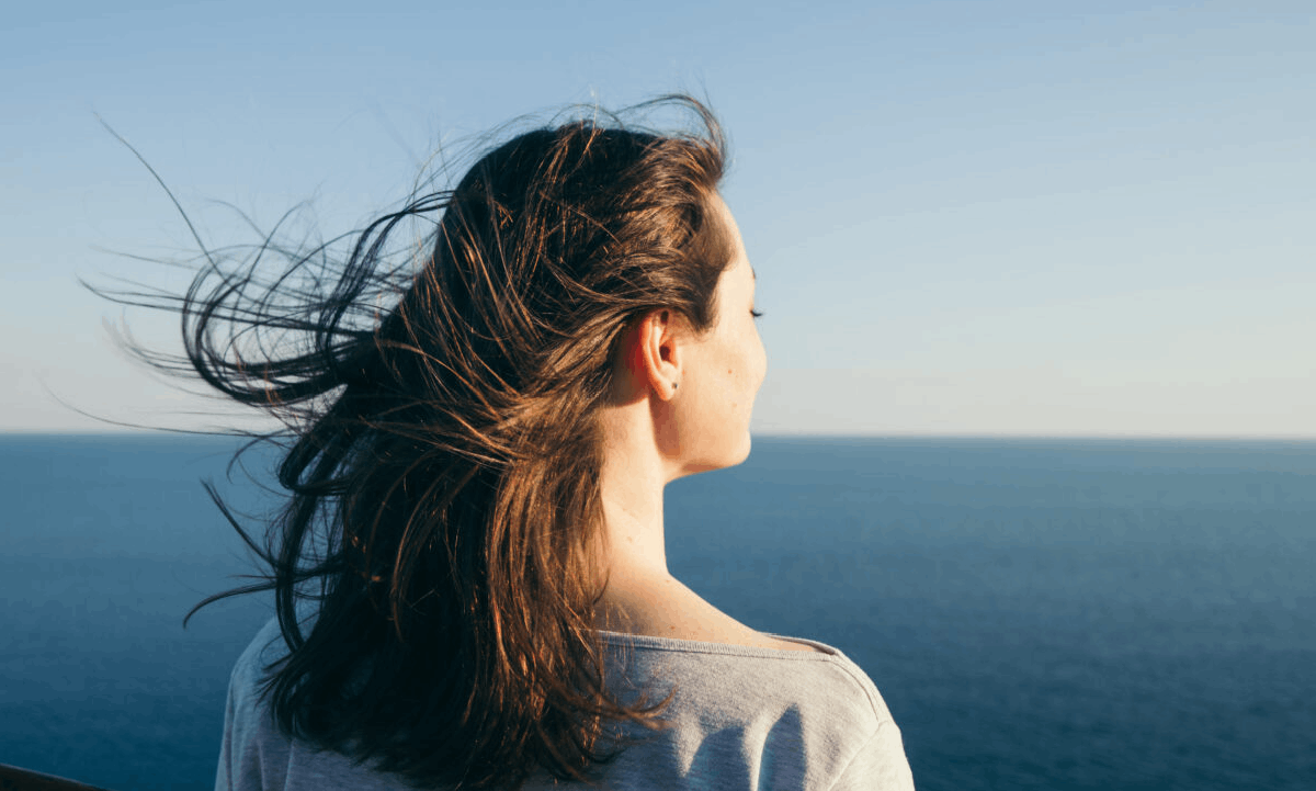 The Cremation Process: A Simple Guide A woman looking out over the ocean, with her hair blowing in the wind beneath a clear blue sky.